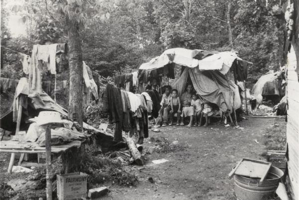 Woman and four children seated in front of a makeshift tent that serves as their home. There is a washtub and washboard in the foreground and laundry hangs from lines tied to trees in the clearing.