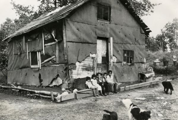Five children seated in front of a dilapidated shack that serves as a dwelling. Three dogs roam in the front yard.