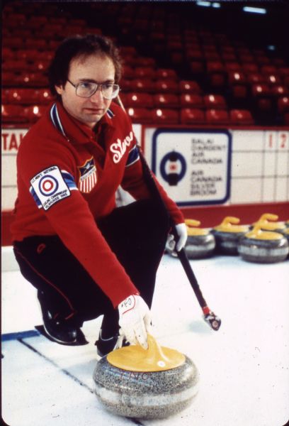 Steve Brown of Madison crouching on ice and holding a curling stone. He wears sweater with a patch that reads: "U.S. Men's National Curling Championship, Milwaukee 1985."