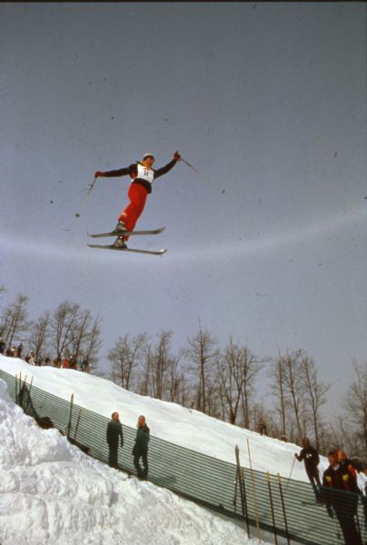 A skier, wearing #34, soars off of a jump with onlookers behind a fence below.