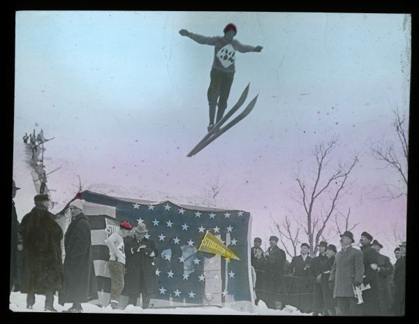 Hand-colored lantern slide view of a ski jumper wearing #32 sailing off of the jump at Stoughton. Spectators watch from the base of the jump which is decorated with a U.S. flag.