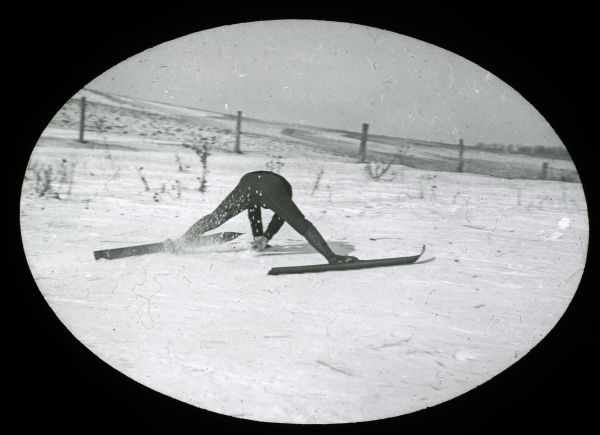 Ski jumper eases to a stop with his legs splayed wide apart, and bent over forwards with their hands touching the ground for balance.