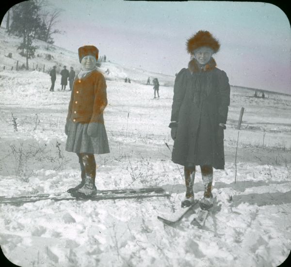 Hand-colored lantern slide of two girls on skis. Other people are in the background.