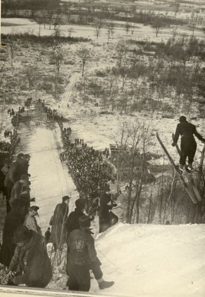 View looking downhill towards a ski jumper in mid-air, with spectators watching from both sides of the landing area below.