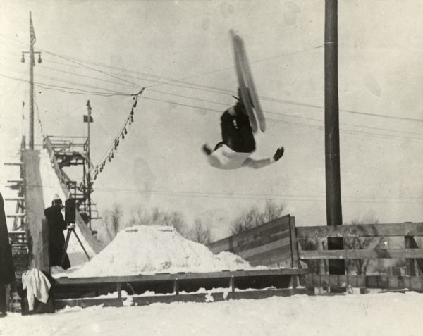 Skier performing a stunt off of a ski jump with a photographer documenting the flip in the background from the base of the jump.