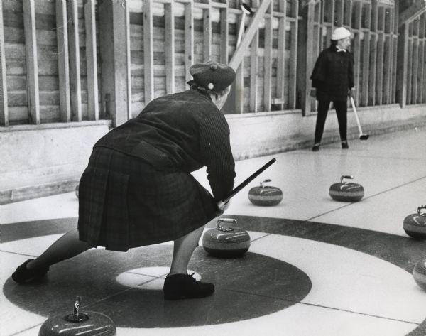 Woman from a visiting curling team from Scotland crouching over the house during a curling match while another woman watches.