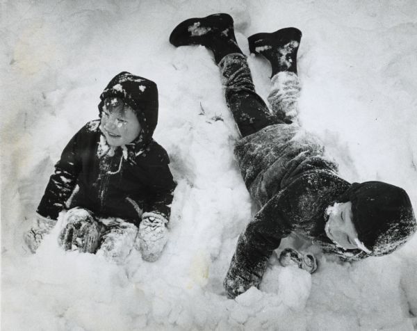 Winter scene with two young boys playing in the snow in front of their home. <i>Milwaukee Journal</i> January 12, 1963 original caption: "The same snow that caused wheels to spin and automobiles to slide throughout Wisconsin also offered a chance for winter fun. Scott Okray, 7, and his brother, Brad, 4, belly flopped and played in the snow Saturday in front of their home, 3244 N. 26th St. Milwaukee reported a four inch snowfall by midmorning. Temperatures are expected to drop sharply by Sunday morning, in the wake of the state's biggest snowstorm of the season."