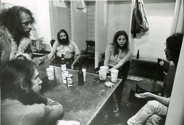 Ian Underwood, Don Preston, Howard Kayland, Gail Zappa, and Frank Zappa discuss science around a backstage table at the Milwaukee Arena following a show by Frank Zappa and the Mothers of Invention. Bottles of Budweiser, cans of 7-Up and Pepsi, and a pack of cigarettes sit on the table.