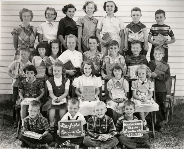 Outdoor group portrait of first through eighth grade students. Most of the students hold books. One holds a chalk board reading Garfield [School], Racine Co., Wis. Another holds a chalk board reading Mrs. Sorenson, Teacher Grades 1-8, 1953-1954.