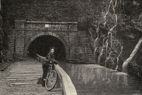 Engraving, probably after a photograph, showing Frank Lenz posing with his safety bicycle near the stone arch entrance of a canal tunnel cut through a mountain. Lenz leans agains a railing along the tow path.