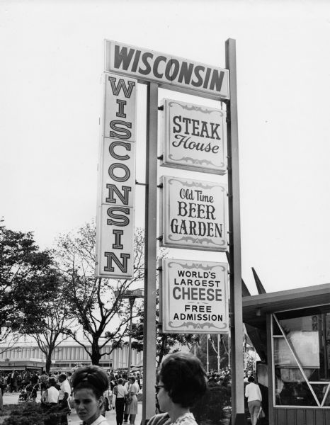 Sign outside the Wisconsin Pavilion at the 1964 World's Fair advertising a steak house, a beer garden, and the world's largest cheese. A portion of the pavilion can be seen at right. Several fair-goers can be seen in the background in front of the General Motors building.
