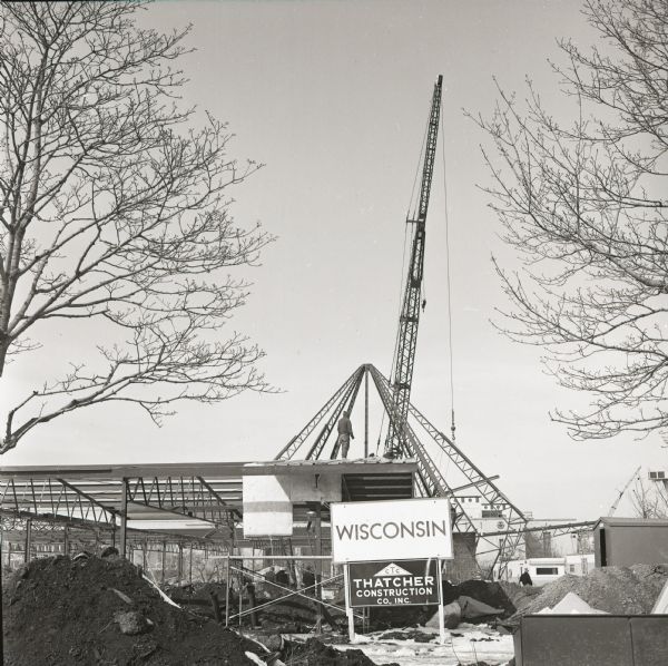 View of the Wisconsin Pavilion at the 1964 World's Fair under construction. The steel frame of the building is visible behind a sign for Thatcher Construction Co. A crane hoists a girder as a man stands atop the structure. Construction trailers can be seen at lower right.