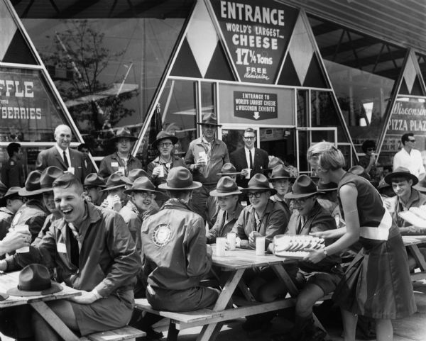 A group of scouts enjoy a snack at picnic tables outside the Wisconsin Pavilion at the 1964 World's Fair. Each scout has a glass of milk and a young woman is offering food from a tray. A sign on the wall advertises the World's Largest Cheese attraction inside the pavilion.
A patch on the back of one scout's shirt reads: "Scout Service Corps, New York World's Fair, 1964-65."