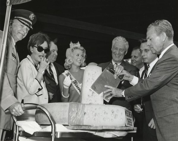 Governor Warren Knowles helps to slice a wedge of cheese. Left to right: an unidentified Colonel; Dorothy Guidry Knowles; State Senator Gerald Lorge; Miss Wisconsin, Sharon Singstock; Governor Knowles; State Senator Taylor Benson; State Senator Martin Schreiber; and World's Fair Vice President, retired Major General William E. Potter.