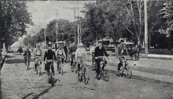 Frank Lenz entering Minneapolis escorted by members of the St. Paul Cycle Club, the Minneapolis Business Men's Bicycle Association, and the Minnesota Division of the League of American Wheelmen. The group rides on a street lined with trees and telephone or telegraph poles.