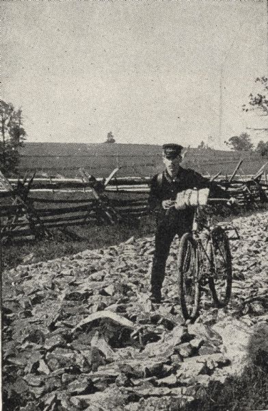 Frank Lenz posed with his bicycle on a badly neglected stretch of road. A split-rail fence encloses a field behind Lenz.
