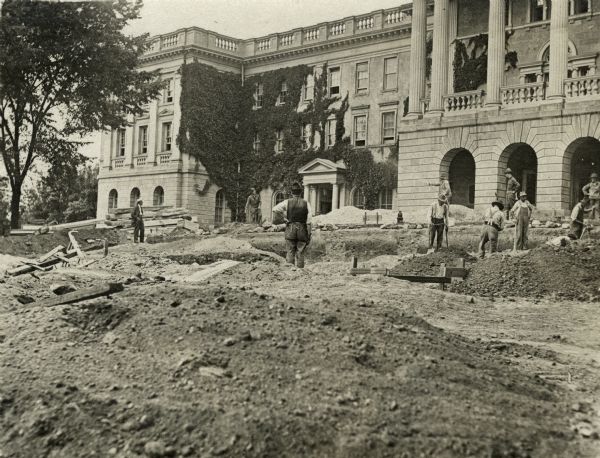 Workers at the site of two graves discovered on Bascom Hill in front of Main Hall (now Bascom Hall). One man sprays water from a hose. The disturbed soil of the two burials can be seen near the center of the image. Madison's first cemetery was located at this site.