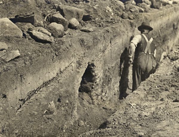 Man with a shovel standing in a ditch in front of two graves discovered on Bascom Hill during preparation to place the Abraham Lincoln monument circle. The hill was the site of the first cemetery in Madison.