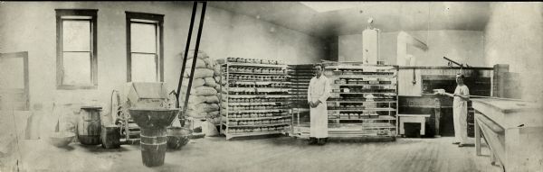 Wide angle view of the interior of a bakery, with one man standing by the oven on the right and another man standing near the center between racks of bread.