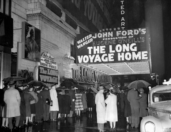 View down sidewalk towards people standing in a line outside a movie theater showing the film <i>The Long Voyage Home</i>. Many people are holding umbrellas. The marquee has the title of the film along with the names of Walter Wanger, John Ford, and Eugene O'Neill.