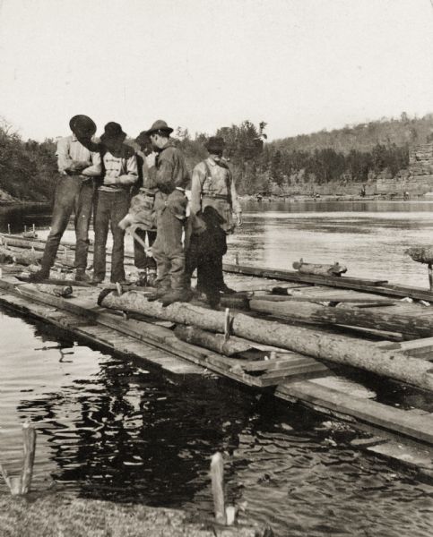 Stereograph of five men standing on a log and a plank of wood while another man is chopping at the log with an axe.