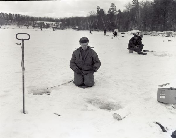 Steve Meleski kneeling near his ice fishing hole at Thunder lake. Other people are fishing on the lake behind him. The shoreline is wooded, and there are buildings in the far background.