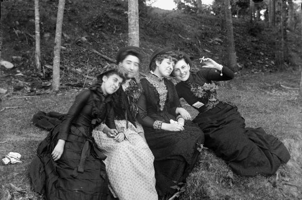 Four women relax and have fun together, sitting on the grass, wearing hats and long dresses. Two of them hold a cigarette in their hand, the other two have a cigarette in their mouth.