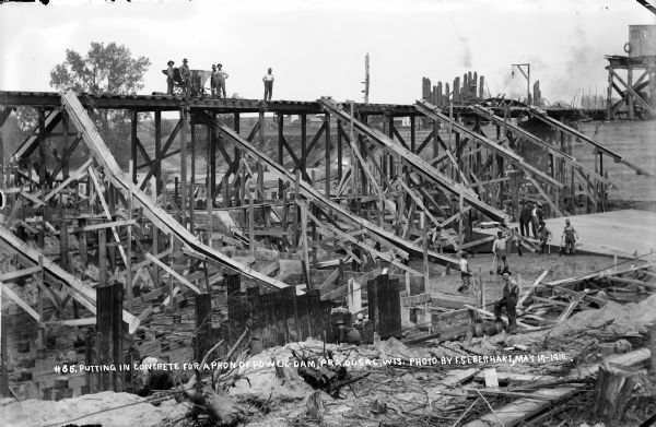 Elevated view of workers putting in concrete to create an apron for the power dam they're building. Two men in suits are standing on a platform on the far right. Across the river are farm buildings.