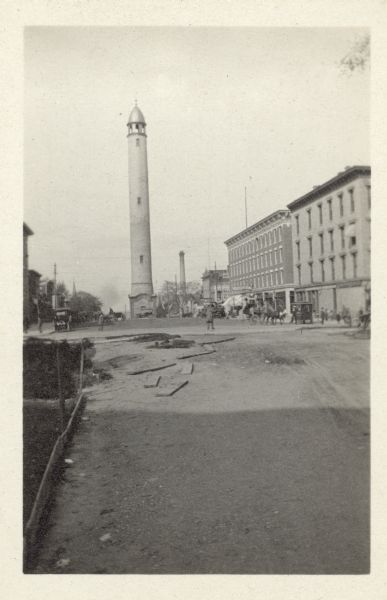 View looking east towards a water tower on East Washington Avenue. There are commercial buildings along the sidewalks on the left and right, and automobiles, horse-drawn vehicles and pedestrians are on the street. A tall smokestack rises above the trees on the right.