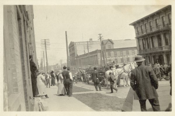 View from sidewalk of people watching a parade coming up the street. A large horse-drawn vehicle is at the front. In the background is a large brick building which has "Madison Saddlery" painted on its side near the rooftop. A bicycle is leaning against the brick building along the left side.