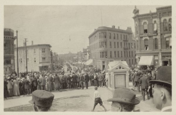 View from the Capitol Square towards a parade coming up State Street. Crowds line the streets. On the left street corner is a building with a sign for the "Madison Medical College."
