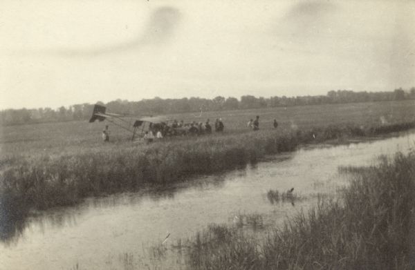 Elevated view across stream in marshy area towards a group of people gathered around an aircraft wreck.