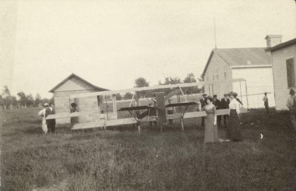 Group of men and women standing around an airplane parked near three buildings in a field.