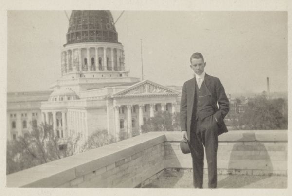 Man in suit, holding a hat, posing on a roof across the street from the Wisconsin State Capitol, where the dome is under construction. In the far background on the right is a smokestack, probably near East Washington Avenue. View appears to be looking north, towards the South Pediment.