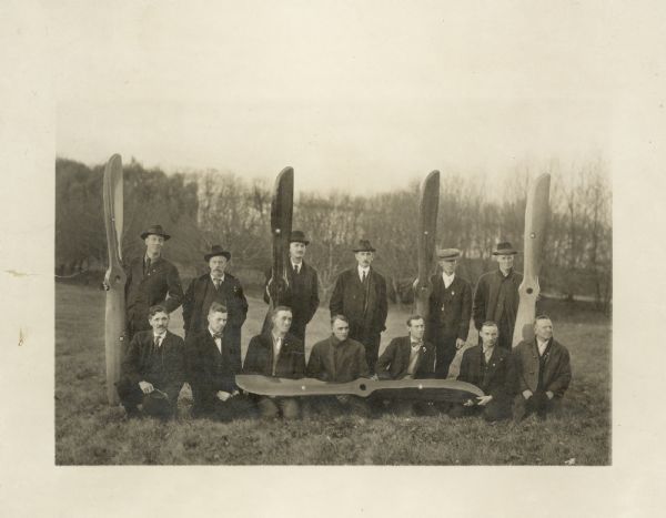 Outdoor group portrait of men from the Forest Products Lab posed along with wooden propellers. They are posed on a hill, with trees in the background.