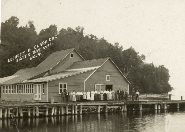 View across water towards a group of people, men, woman, and young girls and boys, posing on a pier in front of a building on the shoreline of Sister Bay. The sign on the front of the building reads: "The Everett B. Clark Company, Seedsmen, Home Office, Milford, Conn." A rocky shoreline and trees are in the background. Caption reads: "Everett B. Clark Co., Sister Bay, Wis."