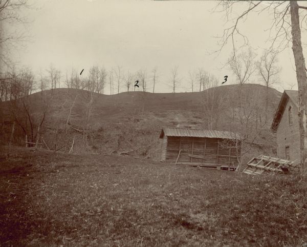 View looking uphill towards three platform mounds. The numbers 1, 2, and 3 are written on the photograph in ink. Part of the way up the hill are a small shed-like building, and a large brick building.