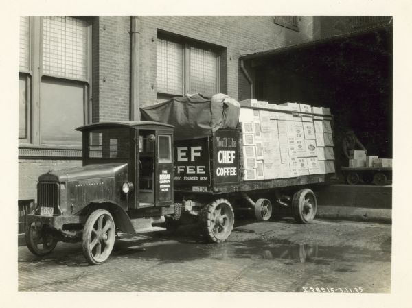 An International Model 63 truck owned by the Berdan Company parked in front of a loading dock. A man on the loading dock is loading boxes of groceries onto the truck's bed.