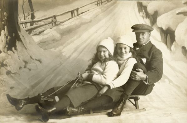 Harold McCormick and his two young daughters pose on a sled/sledge in front of a painted background.