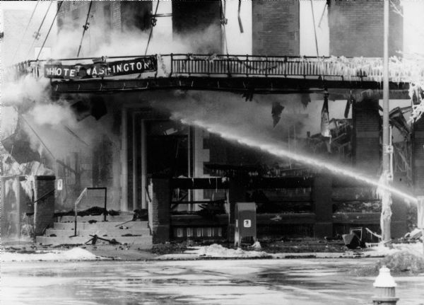 A stream of water shoots into the front entrance of the burning, smoking Hotel Washington. The crumpled hotel sign still hangs on the balcony.