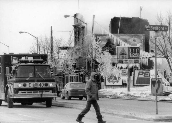 A view from Bedford Street of the smouldering ruins of the Hotel Washington. A pedestrian crosses the street in front of a parked firetruck. Water being used to fight the fire has frozen into long icicles on signs and power lines.