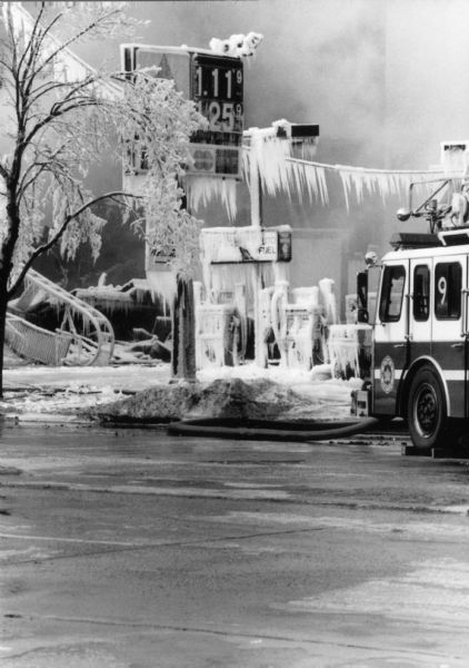Water from firehoses has frozen into ice that coats power wires, trees, and pumps at the gas station next to the Hotel Washington, which burns in the background. A fire truck is parked at the gas station. Ruins of the hotel are barely visible through smoke.