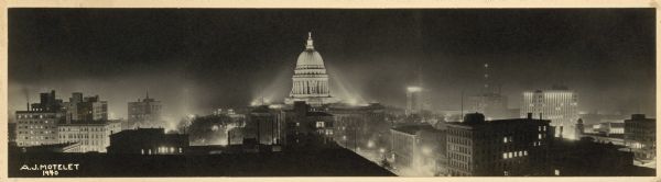 Panorama view, elevated, of the Wisconsin State Capitol at night from the roof of a building on Monona Avenue. The First National Bank and the Belmont Hotel are on the right. On the left is the Park Hotel and Grace Episcopal Church at the top of West Washington Avenue.