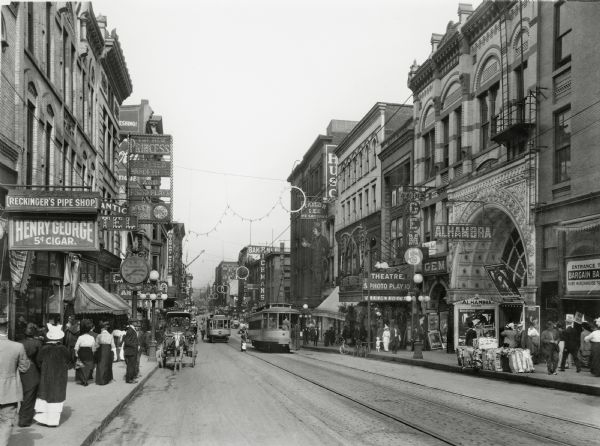 Seventh Street looking east. Streetcars are running in both diretions, and horse-drawn vehicles and bicycles are near the curb. The sidewalks are filled with pedestrians. The Alhambra Theater is on the right, with a poster for "Broken Nose Bailey" displayed in the arch. The New Princess Refined Vaudeville and Photo-Plays theater is on the left.