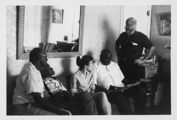 Group of people sitting on a sofa, with one man standing on the right. The man sitting on the far right holding papers in his hands is Amzie Moore, noted civil rights activist and entrepreneur.