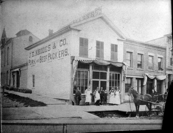 View from street of the front of the J.E. Rhodes & Co., Pork and Beef Packers building on Main Street. A group of men, with a cat and a dog, are posing in front of the entrance. There is a horse and buggy at the curb. The steeple of a church is visible above rooftops on the left.