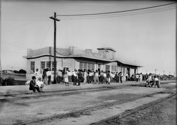 View of a crowd standing on platform near the tracks at Harlingen Railroad Depot, awaiting a train's arrival. A sign posted on the platform is written in English and Spanish.