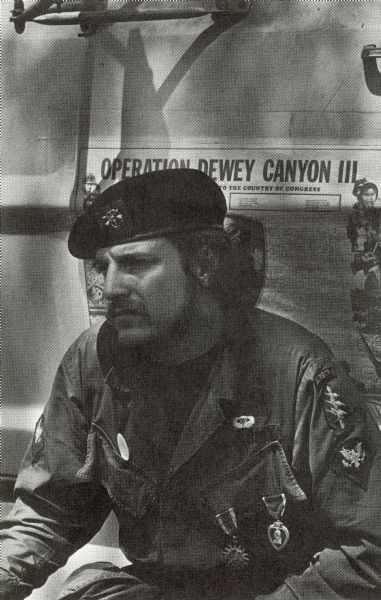 A Vietnam veteran with a moustache wearing a beret and military medals and patches on his shirt. He is at an anti-war demonstration and took part in the Winter Soldier Hearings. A newspaper article posted behind him has the headline: "Operation Dewey Canyon III."