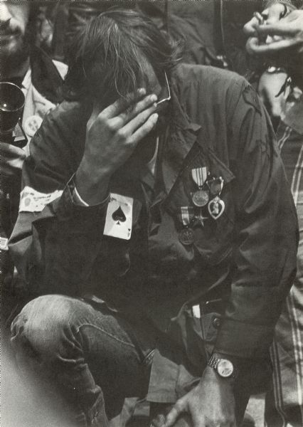 A Vietnam veteran is kneeling on the ground with his hand shielding his face. There are medals and an Ace of Spades playing card pinned to his shirt. He is at an anti-war demonstration and took part in the Winter Soldier Hearings. There are people behind him, and the man on the left is holding a camera.