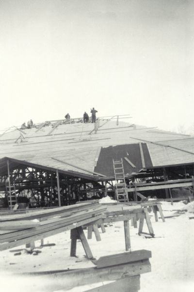 View from snowy ground of men working on the roof. In the foreground are stacks of wood on sawhorses.